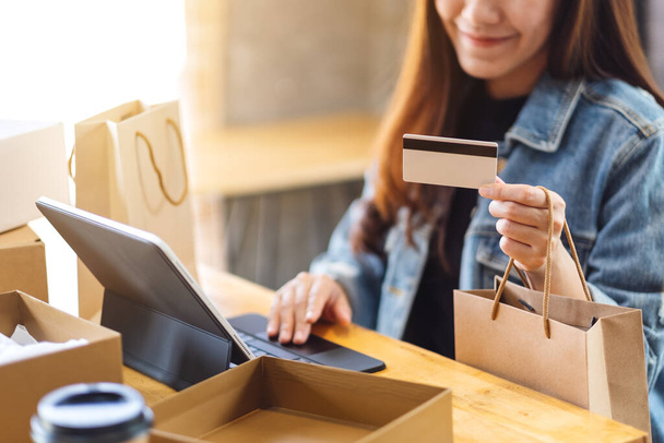 Nahaufnahme einer jungen asiatischen Frau mit Tablet-PC und Kreditkarte beim Online-Einkauf mit Postpaketkasten und Einkaufstaschen auf dem Tisch - Foto, Bild