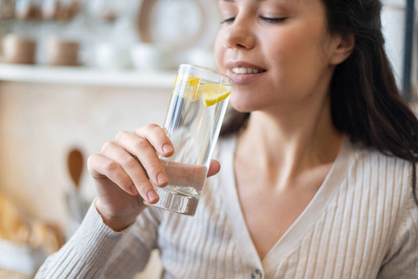 Refreshment concept. Young woman drinking water with lemon, holding glass, standing in kitchen, closeup - Foto, imagen