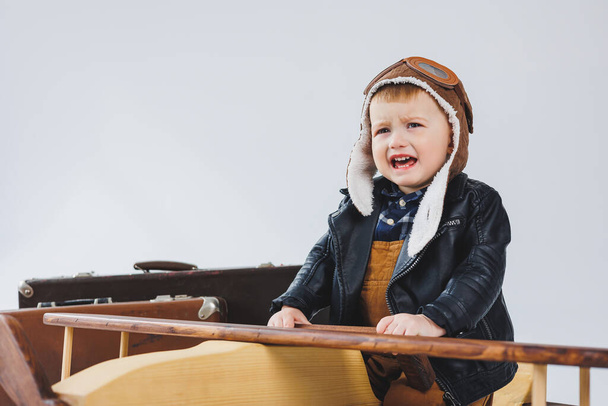 El niño está llorando con una chaqueta de cuero y un sombrero de piloto, un avión de madera, maletas marrones. Juguetes de madera para niños. Emociones del bebé - Foto, imagen