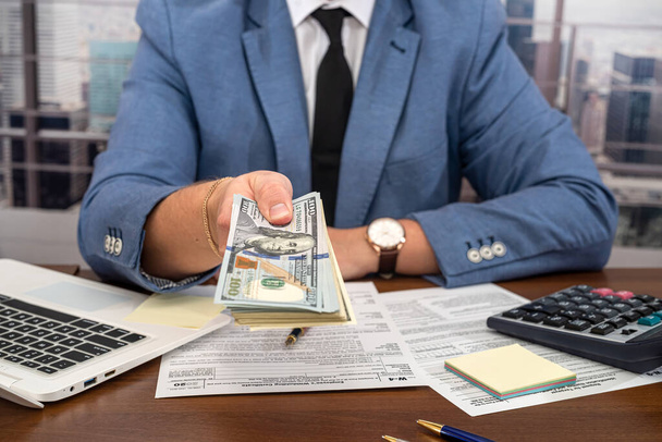 young businessman in a black stylish suit at a spacious table counts dollars and fills out forms. The concept of tax forms and dollars - Photo, Image