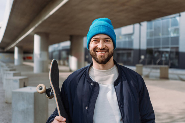 Trendy stoere mannen portret nam om de camera, het houden van skate board en het dragen van pet. Skateboard spot.  - Foto, afbeelding