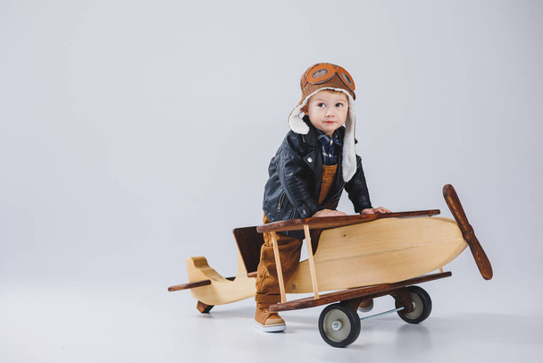 U dřevěného letadla stojí šťastný chlapec v helmě a v kabátě pilota. Portrét dětského pilota, dítě v kožené bundě. Dřevěné hračky. Eko letadlo ze stromu - Fotografie, Obrázek