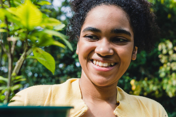 primer plano retrato joven negro chica con afro al aire libre en el campo sonriendo en un día soleado, mirando a una planta que ella va a trasplante. - Foto, Imagen