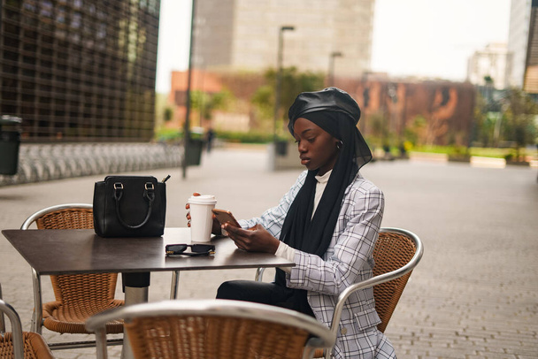Σοβαρή μαύρη γυναίκα που χρησιμοποιεί smartphone στο τραπέζι σε υπαίθριο καφέ - Φωτογραφία, εικόνα