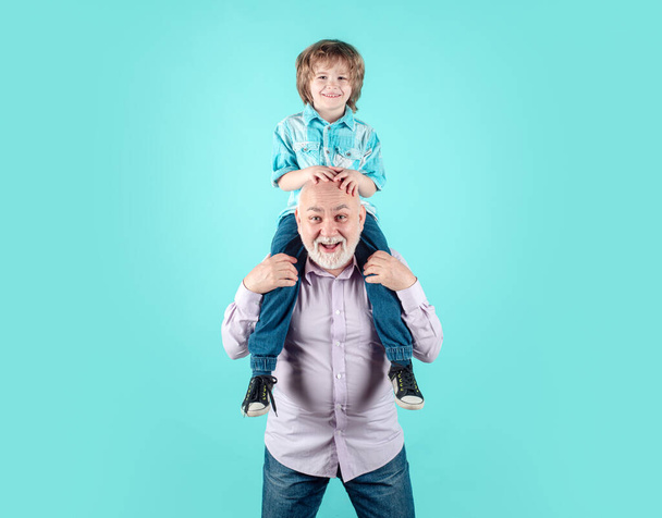 Старый дед и маленький ребенок внук на спине со смешным лицом, изолированным на синем в студии. Дедушка и милый мальчик с забавным лицом. - Фото, изображение
