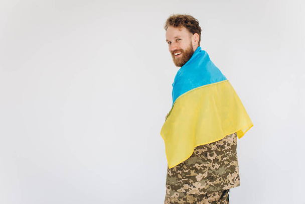 Feliz soldado patriota ucraniano em uniforme militar segurando uma bandeira amarela e azul em um fundo branco - Foto, Imagem