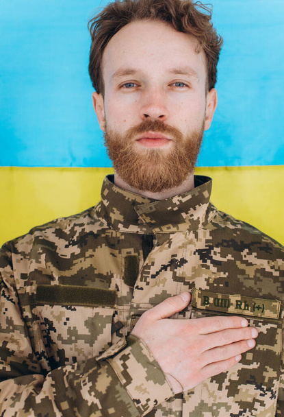 Askeri üniformalı Ukraynalı vatansever asker, sarı ve mavi bir bayrağın arka planında bir kalbe el kaldırdı. - Fotoğraf, Görsel
