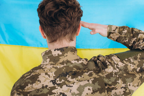 制服を着たウクライナの愛国者兵士は、黄色と青の旗の背景を背景に彼の頭に手を保持しています - 写真・画像