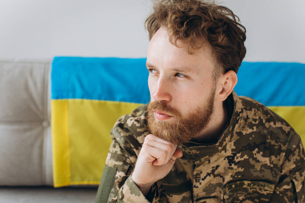 Porträt eines emotionalen jungen ukrainischen Patrioten in Militäruniform, der auf einem Sofa mit gelb-blauer Flagge sitzt. - Foto, Bild
