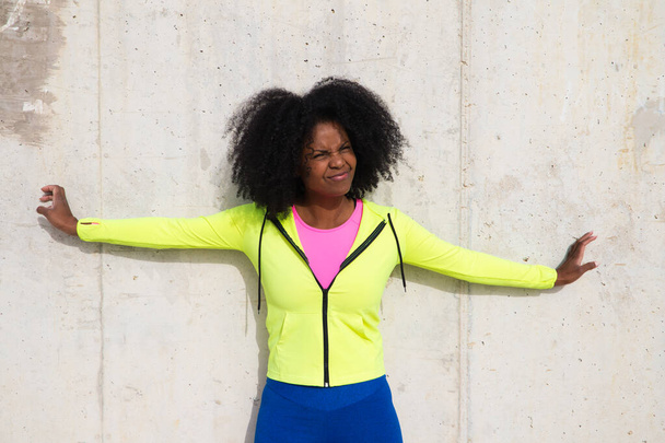 Beautiful young Afro-American woman in bright green and pink sportswear on a grey concrete wall texture background. Woman makes different expressions. Laughing, serious, happy, sad, thinking - Photo, Image