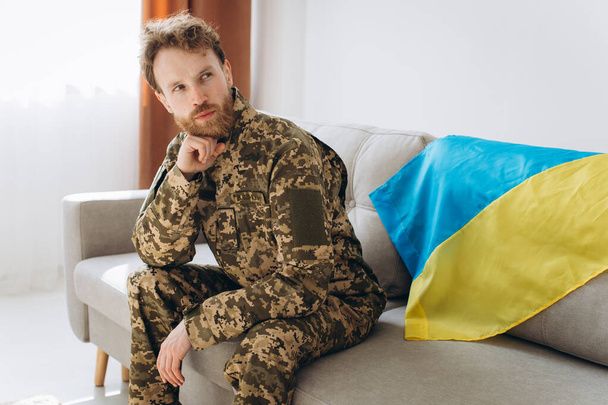 Retrato de un joven soldado patriota ucraniano con uniforme militar sentado en la oficina en un sofá con una bandera amarilla y azul. - Foto, Imagen