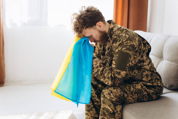 Portrét mladého emocionálního ukrajinského patriota ve vojenské uniformě sedícího na gauči na kanclu se žlutou a modrou vlajkou. - Fotografie, Obrázek