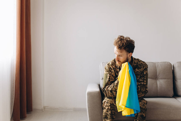 Muotokuva emotionaalinen nuori ukrainalainen patriootti sotilas sotilasunivormussa istuu toimistossa sohvalla pitäen keltainen ja sininen lippu. - Valokuva, kuva