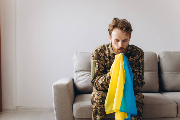 Portrait of an emotional young Ukrainian patriot soldier in military uniform sitting on the office on the couch holding a yellow and blue flag. - Fotoğraf, Görsel