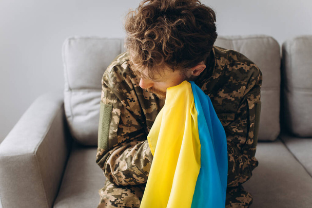 Retrato de um jovem soldado patriota ucraniano emocional em uniforme militar sentado no escritório no sofá segurando uma bandeira amarela e azul. - Foto, Imagem
