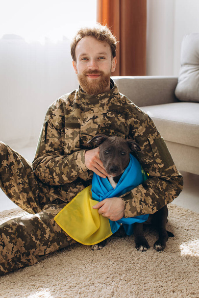 Soldat ukrainien étreignant et enveloppant le drapeau ukrainien de son fidèle ami autour d'un chien Amstaff dans le bureau - Photo, image