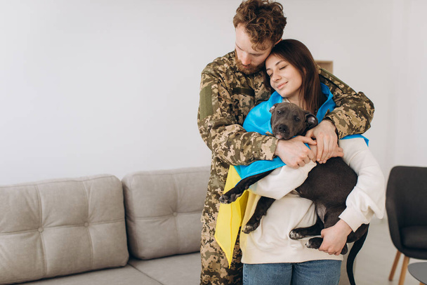 Um casal ucraniano, um soldado de uniforme militar e uma menina envolto em uma bandeira ucraniana segurar um cão em seus braços, felizes juntos. - Foto, Imagem