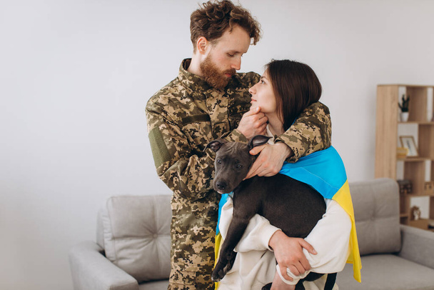 Ένα ζευγάρι Ουκρανών, ένας στρατιώτης με στρατιωτική στολή και ένα κορίτσι τυλιγμένο σε μια ουκρανική σημαία κρατούν ένα σκύλο στην αγκαλιά τους, ευτυχισμένοι μαζί.. - Φωτογραφία, εικόνα