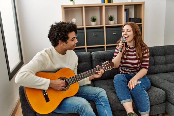 Νεαρό ζευγάρι παίζει κλασική κιθάρα και τραγουδά χρησιμοποιώντας μικρόφωνο στο σπίτι. - Φωτογραφία, εικόνα
