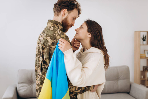 A Ukrainian girl hugs and holds a yellow and blue flag of a military man in uniform and says goodbye to him. - Foto, Imagen