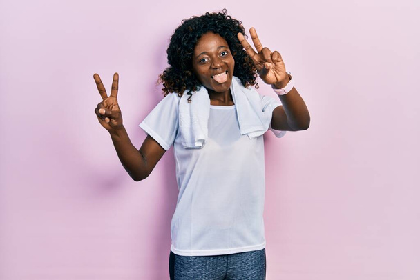 Νεαρή Αφροαμερικανή που φοράει αθλητικά και πετσέτα χαμογελώντας με τη γλώσσα έξω δείχνοντας τα δάχτυλα και των δύο χεριών να κάνουν το σύμβολο της νίκης. Νούμερο δύο..  - Φωτογραφία, εικόνα