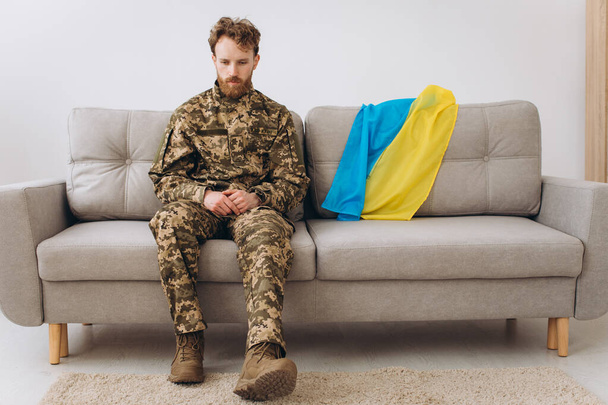 Портрет эмоционального молодого украинского патриота в военной форме, сидящего в офисе на диване с желтым и синим флагом. - Фото, изображение