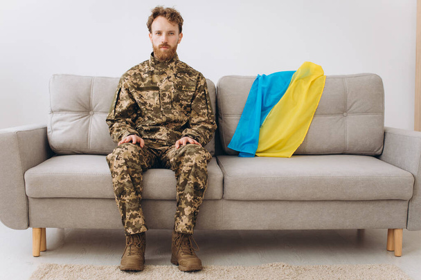 Portrait of an emotional young Ukrainian patriot soldier in military uniform sitting on the office on a sofa with a yellow and blue flag. - Photo, Image