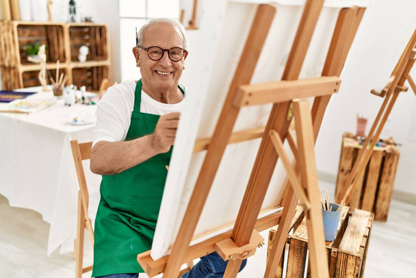 Ανώτερος γκριζομάλλης καλλιτέχνης χαμογελά χαρούμενος ζωγραφική στο στούντιο τέχνης. - Φωτογραφία, εικόνα