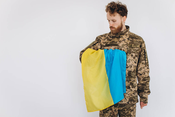 Πορτρέτο ενός συναισθηματικού νεαρού γενειοφόρου Ουκρανού πατριώτη στρατιώτη με στρατιωτική στολή που κρατά σημαία στο γραφείο - Φωτογραφία, εικόνα