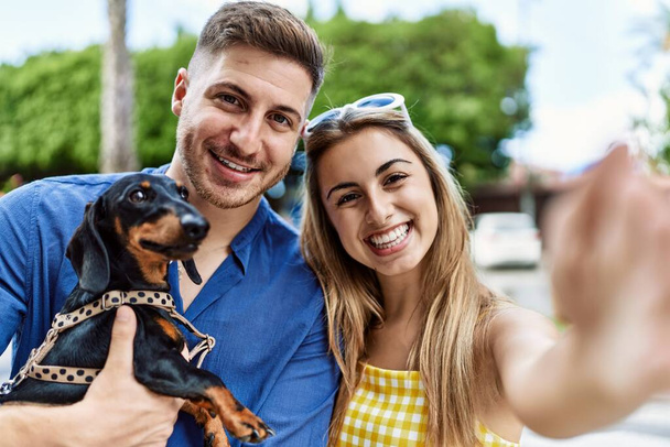 joven caucásico pareja sonriendo feliz hacer selfie por la cámara con perro en la ciudad. - Foto, imagen