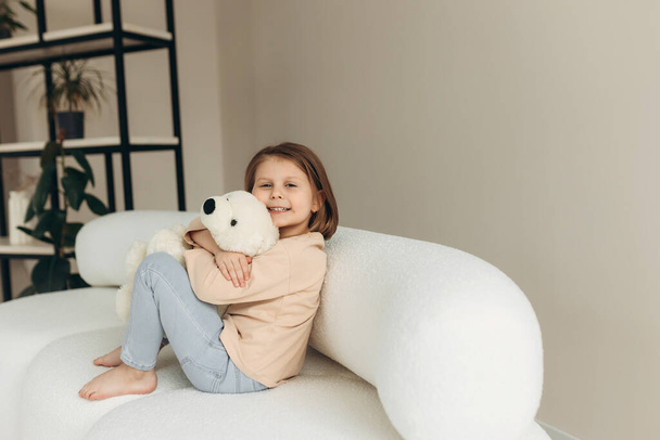 Χαριτωμένο κοριτσάκι αγκαλιάζει ένα πολικό αρκουδάκι, κοιτάζοντας την κάμερα και χαμογελώντας ενώ κάθεται στο σπίτι στο κρεβάτι - Φωτογραφία, εικόνα