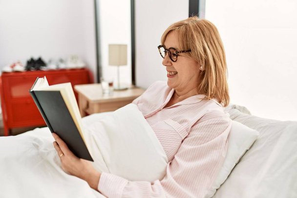 Middelbare leeftijd blonde vrouw lezen boek liggend op het bed thuis. - Foto, afbeelding
