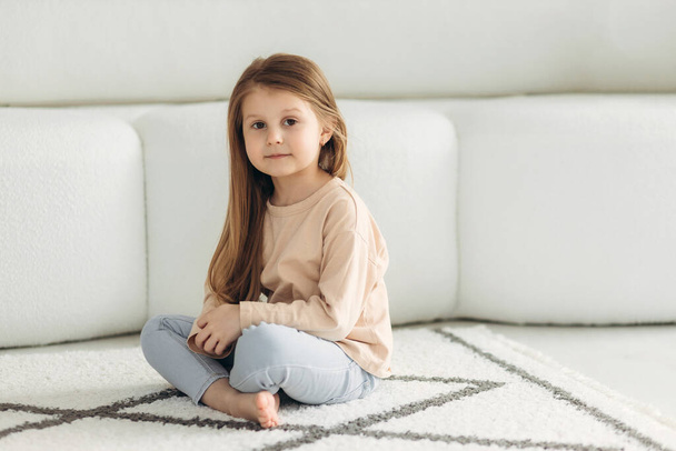 Portrét malé roztomilé holčičky. Sedí na podlaze u pohovky. Dívka se dívá do kamery - Fotografie, Obrázek