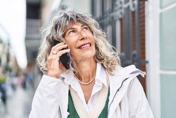 Kobieta w średnim wieku uśmiecha się pewnie rozmawiając na smartfonie na ulicy - Zdjęcie, obraz
