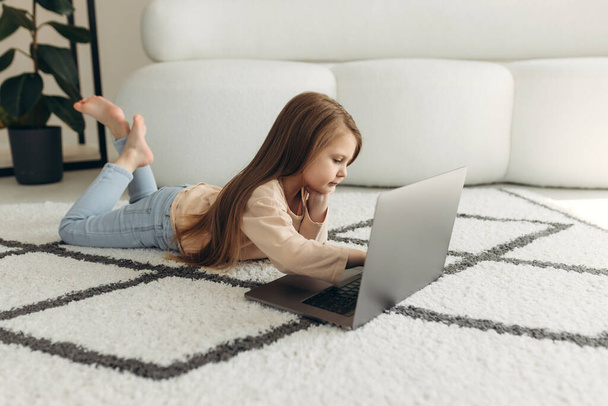 Cute baby looking at laptop screen. Little girl lying at home on the couch using the Internet. Dependence on technology or computer. Online education. - 写真・画像