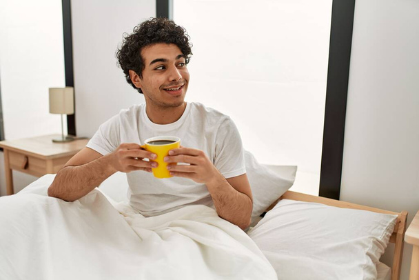 Jovem hispânico bebendo café sentado na cama no quarto. - Foto, Imagem