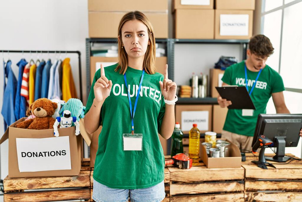Young blonde girl wearing volunteer t shirt at donation stand pointing up looking sad and upset, indicating direction with fingers, unhappy and depressed.  - Photo, Image