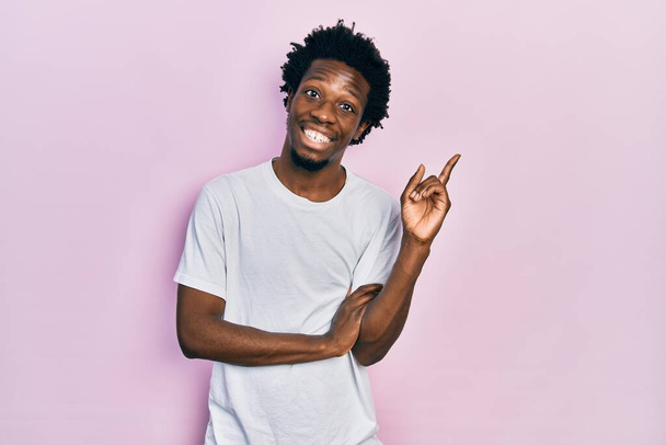 Νεαρός Αφροαμερικάνος που φοράει λευκό μπλουζάκι με μεγάλο χαμόγελο στο πρόσωπο, δείχνοντας με το χέρι και το δάχτυλο στο πλάι κοιτάζοντας την κάμερα.  - Φωτογραφία, εικόνα