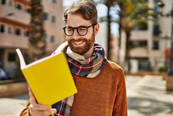 Νεαρός καυκάσιος με γενειάδα φοράει γυαλιά διαβάζοντας ένα βιβλίο σε μια ηλιόλουστη μέρα - Φωτογραφία, εικόνα