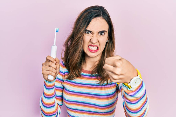 Young hispanic girl holding electric toothbrush annoyed and frustrated shouting with anger, yelling crazy with anger and hand raised  - Foto, Bild