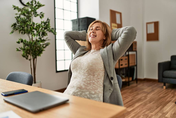 Bizneswoman w średnim wieku uśmiechnięta szczęśliwa zrelaksowana z rękami na głowie w biurze. - Zdjęcie, obraz