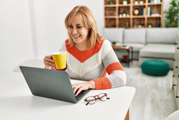 Блондинка середнього віку працює з ноутбуком і п'є каву вдома
. - Фото, зображення