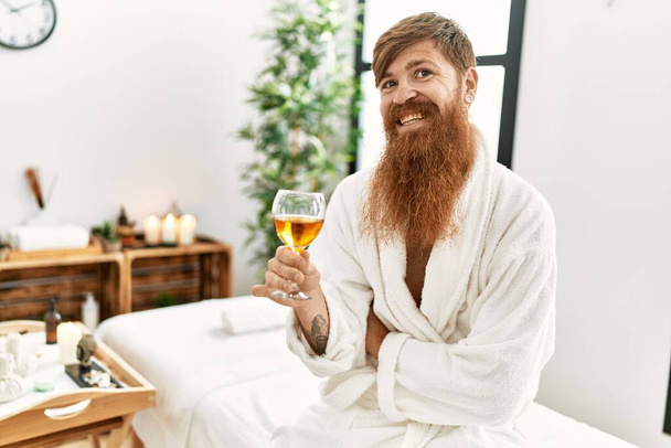 Young redhead man wearing bathrobe drinking champagne at beauty center - Фото, изображение