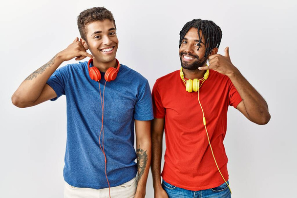Young hispanic brothers standing over isolated background wearing headphones smiling doing phone gesture with hand and fingers like talking on the telephone. communicating concepts.  - Φωτογραφία, εικόνα