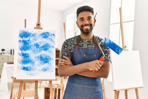 młody afrykański artysta amerykański człowiek uśmiecha się szczęśliwy z ramionami skrzyżowane gest trzymając rolki farby w studio sztuki. - Zdjęcie, obraz