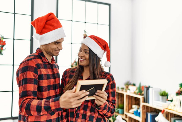 Young latin couple celebrating christmas holding frame at home. - Zdjęcie, obraz