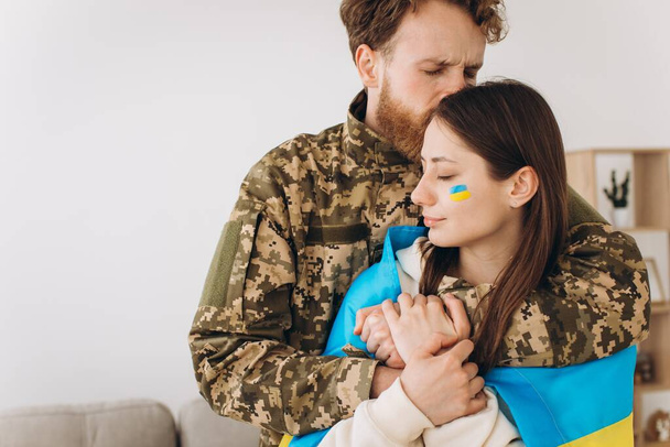 Oekraïense echtpaar, militair in uniform knuffelt en wikkelt zijn vrouw in de Oekraïense vlag. Het begrip patriottisme. - Foto, afbeelding