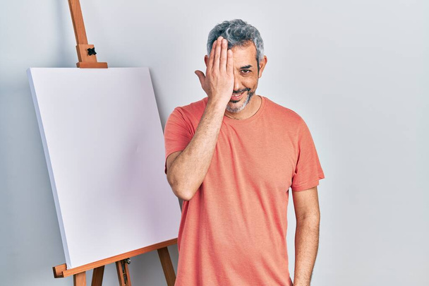Handsome middle age man with grey hair standing by painter easel stand covering one eye with hand, confident smile on face and surprise emotion.  - Foto, imagen