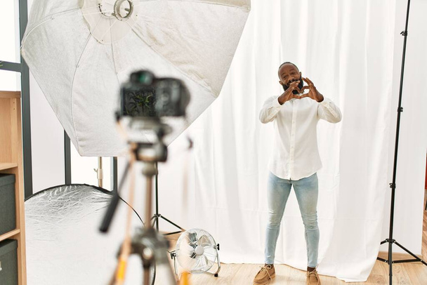 Homme afro-américain se faisant passer pour modèle dans un studio de photographie souriant d'amour montrant le symbole du cœur et la forme avec les mains. concept romantique.  - Photo, image