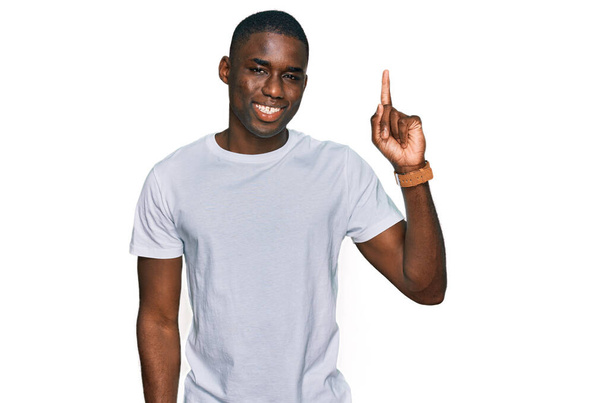 Jeune homme afro-américain portant un t-shirt blanc décontracté pointant du doigt avec une idée réussie. Sorti et heureux. numéro un.  - Photo, image